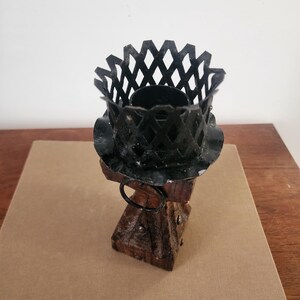 May include: A candle holder featuring a dark brown wooden base with metal studs and a circular ring attachment. The top consists of a black metal lattice cage with a scalloped edge, designed to securely hold a candle in an upright position.