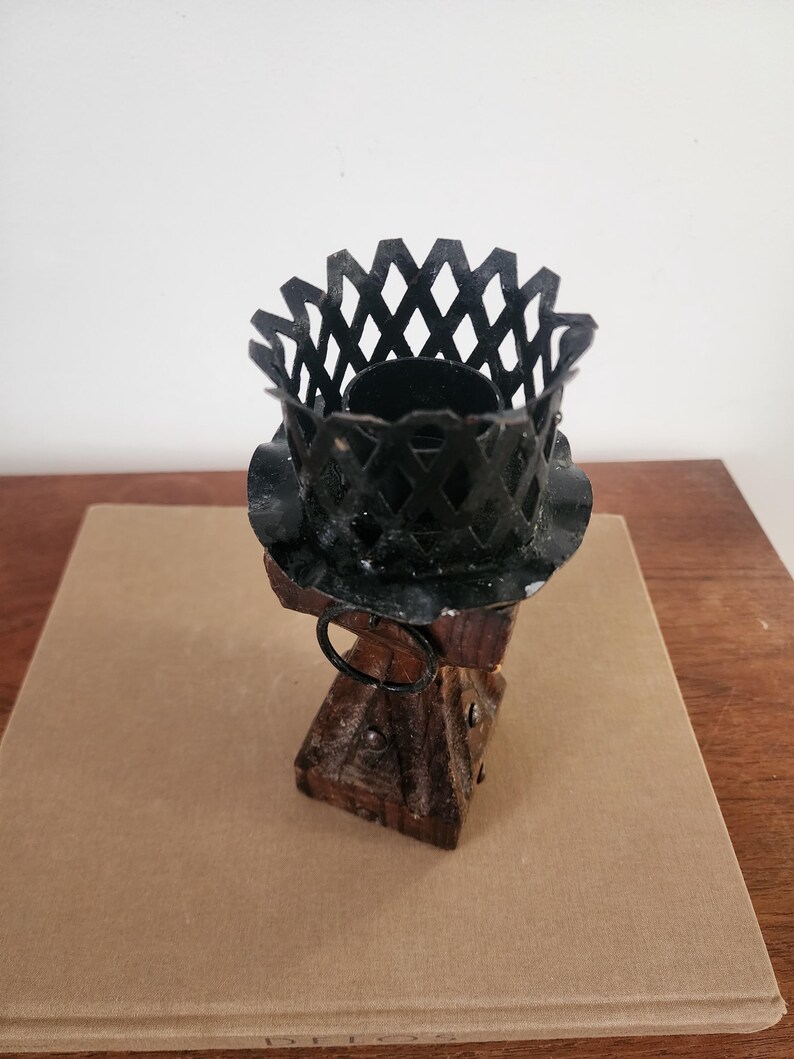 May include: A candle holder featuring a dark brown wooden base with metal studs and a circular ring attachment. The top consists of a black metal lattice cage with a scalloped edge, designed to securely hold a candle in an upright position.