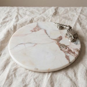 May include: A circular white marble serving board featuring prominent pink and grey veining throughout the stone surface. A decorative silver-toned metal accent shaped like a cluster of seashells is attached to the upper right edge of the board.