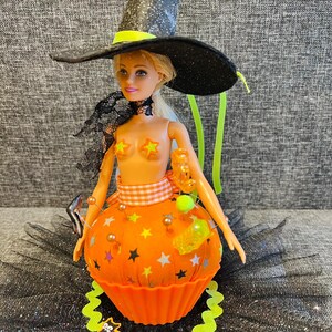 May include: A doll dressed as a Halloween witch, wearing a black glittery hat with a yellow band, and an orange cupcake-shaped dress decorated with stars. The doll has a black lace choker and star-shaped pasties. A neon green necklace and a black cat decoration complete the look.