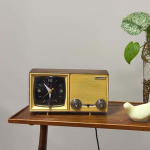 May include: A rectangular yellow and brown clock radio with a black face, white numerals, and silver hands. Beside it sits a pale yellow ceramic bird figurine. The clock radio features a ribbed yellow speaker grille and two circular silver knobs.