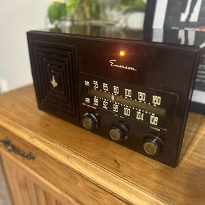 May include: A vintage Emerson radio in a dark brown finish. The radio features a speaker grill, tuning dials, and a frequency display. The word "Emerson" is printed on the front. The radio is placed on a wooden cabinet.