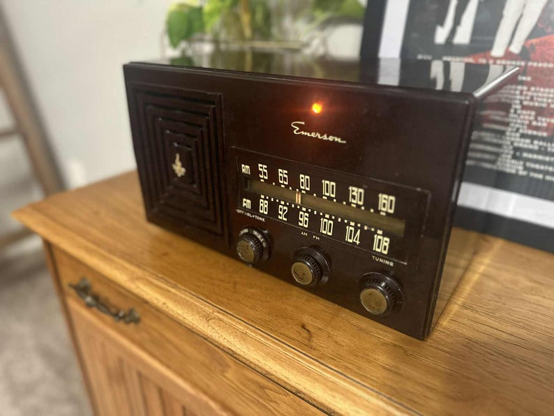 May include: A vintage Emerson radio in a dark brown finish. The radio features a speaker grill, tuning dials, and a frequency display. The word "Emerson" is printed on the front. The radio is placed on a wooden cabinet.