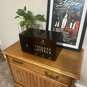 May include: A vintage brown Emerson radio with a speaker grill and tuning dial sits atop a wooden cabinet. A framed poster of "The Strike" North American Tour is behind the radio. The cabinet has two drawers with metal handles.