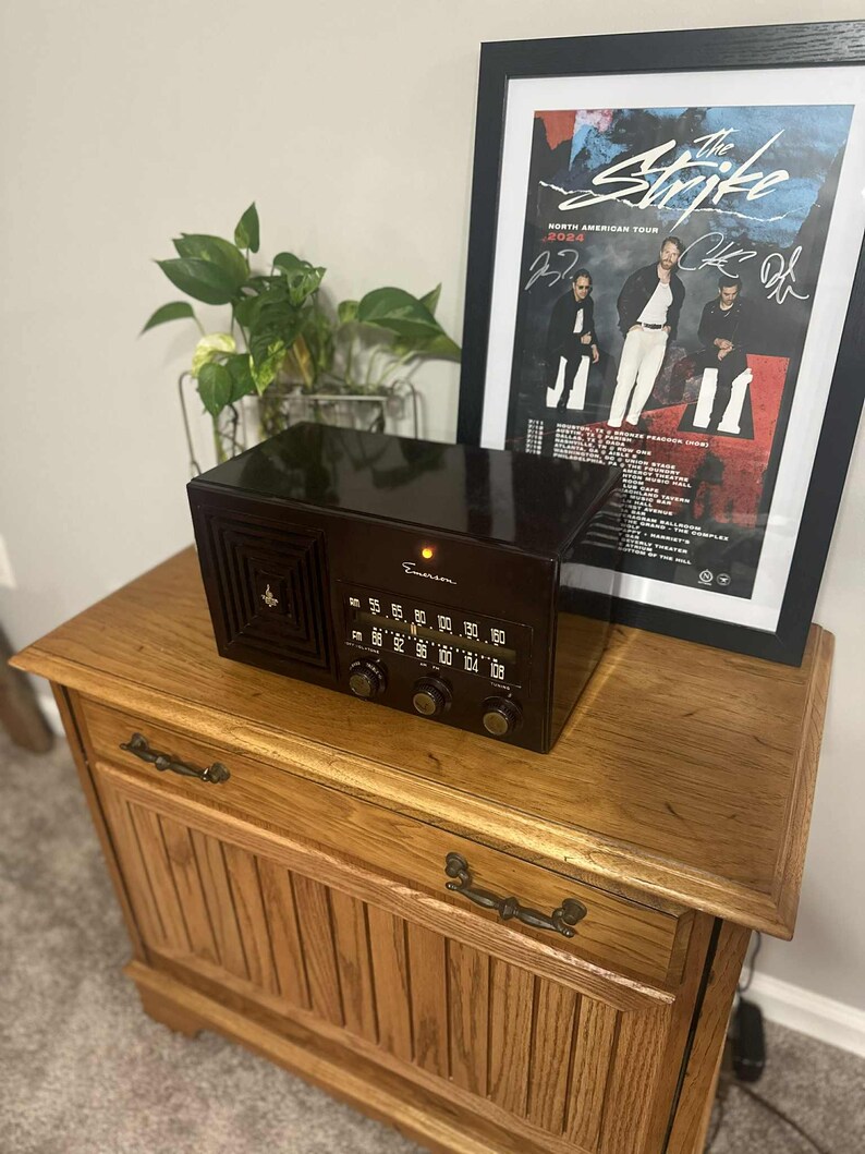 May include: A vintage brown Emerson radio with a speaker grill and tuning dial sits atop a wooden cabinet. A framed poster of "The Strike" North American Tour is behind the radio. The cabinet has two drawers with metal handles.
