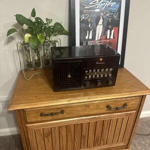 May include: A vintage Emerson radio with a dark brown finish sits atop a wooden cabinet. The cabinet has a drawer and a lower door with vertical paneling. A framed poster and a plant in a glass vase are also on the cabinet.
