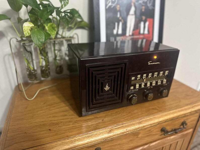 May include: A vintage Emerson radio in a dark brown finish, featuring a square speaker grille and a tuning dial. The radio sits on a wooden cabinet, with a small light illuminated above the dial. A plant in a glass vase is visible in the background.