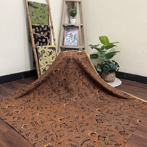 May include: A large, brown fabric with a detailed floral pattern is draped over a wooden surface. Several other patterned fabrics in green, black, and yellow are displayed on a bamboo ladder. A wooden shelf with plants and a framed picture is in the background.