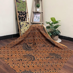 以下が含まれることがあります： スタイリッシュな鳥と花のモチーフが特徴の、詳細な黒い模様が施された大きな茶色のバティック織物。この生地は木製の表面にドレープされており、その複雑なデザインが際立っています。緑、黒、黄色の他のバティック織物は、竹製の梯子に展示されています。