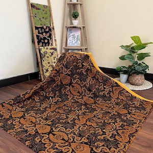 Könnte beinhalten: Ein großes Textil mit einem komplizierten schwarzen und goldenen Muster, das stilisierte Blumen- und abstrakte Formen aufweist. Der Stoff ist über einer Holzoberfläche drapiert, mit einem gelben Rand. Weitere Textilien werden auf einer Bambusleiter ausgestellt.