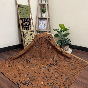 以下が含まれることがあります： 詳細な黒い鳥と花の模様が施された、大きな茶色の織物。木製の表面に生地が掛けられており、複雑なデザインが際立っています。竹の梯子には、花の模様が施された追加の織物が展示されています。