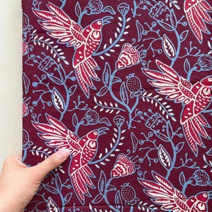Könnte beinhalten: Ein tiefburgunderroter Stoff mit einem sich wiederholenden Muster aus stilisierten fliegenden Vögeln, durchsetzt mit hellblauen Blumenranken, Blättern und glockenförmigen Blüten. Das Design ist in einem flachen, grafischen Stil auf den Stoff gedruckt.