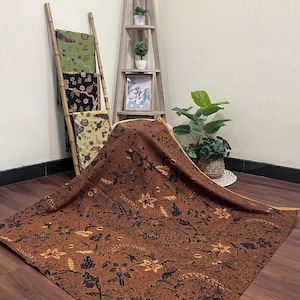 Könnte beinhalten: Ein brauner Stoff mit floralem Muster, der auf einer Holzoberfläche präsentiert wird. Der Stoff weist schwarze und beige florale Designs auf. Weitere Textilien mit ähnlichen Mustern werden auf einer Bambusleiter im Hintergrund ausgestellt.