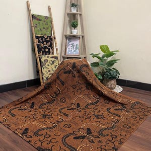 May include: A brown textile with a black and gold dragon pattern is displayed on a wooden floor. The textile is draped over a wooden shelf and a bamboo ladder with other patterned textiles. Decorative plants and a framed picture are also visible.
