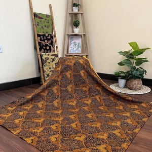 Może przedstawiać: Duży materiał z brązowo-złotym wzorem kwiatowym rozłożony na drewnianej podłodze. Inne wzorzyste tkaniny są eksponowane na bambusowej drabinie. Drewniana półka z roślinami i oprawionym zdjęciem znajduje się w tle.