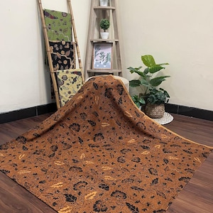 Puede incluir: Una tela marrón grande con un estampado floral negro se extiende sobre una superficie de madera. Otras telas con diseños florales en verde, negro y amarillo se exhiben en una escalera de madera. Una estantería de madera con plantas y una imagen enmarcada está en el fondo.