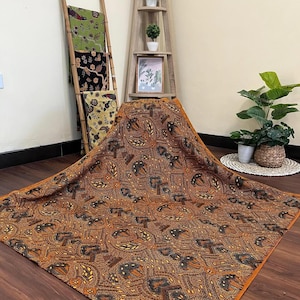 Puede incluir: Una tela grande estampada en tonos marrones y naranjas, con un diseño repetido de formas estilizadas. La tela está drapeada sobre una superficie de madera, con otras telas estampadas visibles en el fondo.