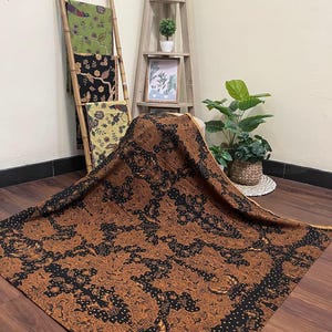Puede incluir: Un gran textil con un estampado floral marrón y negro está drapeado sobre una superficie. Otros textiles con estampados florales en verde, negro y amarillo se exhiben en una escalera de bambú. Una estantería de madera con plantas y un cuadro enmarcado se encuentra en el fondo.