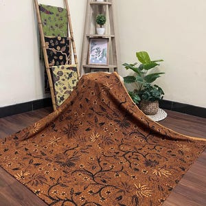 Puede incluir: Una tela marrón con un estampado floral negro está drapeada sobre una superficie. La tela es el punto focal, con un diseño detallado de flores y vides. Otras telas con estampados florales se exhiben en una escalera de bambú en el fondo.