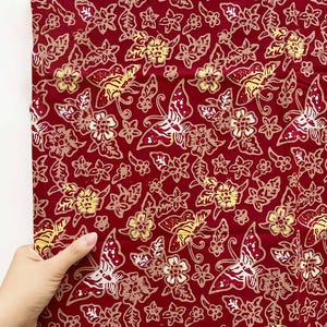 Könnte beinhalten: Ein tiefroter Stoff mit einem sich wiederholenden Batik-Muster aus Schmetterlingen und kleinen Blumen in hellen Beige- und Gelbtönen. Das Textil zeigt ein dichtes, kompliziertes Design mit organischen Formen und feinen Linien auf der Oberfläche.