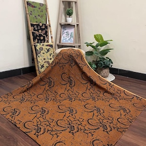 May include: A large, rust-colored fabric with a black floral pattern is spread out on a wooden surface. Three other patterned fabrics are displayed on a bamboo ladder. A wooden shelf with plants and a framed picture is in the background.