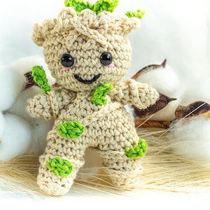 May include: A crocheted doll resembling a character with a light brown body, green accents, and black eyes. The doll has a smiling face and is posed in front of cotton bolls and dried plant material. The doll is approximately 13 cm tall.