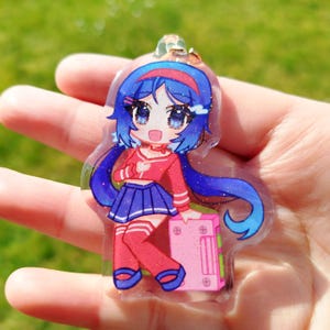 May include: A clear acrylic charm featuring a cartoon anime character with blue hair, wearing a red top, blue skirt, and red knee-high socks. The character is holding a pink box. The charm has a gold-colored metal loop for attachment.