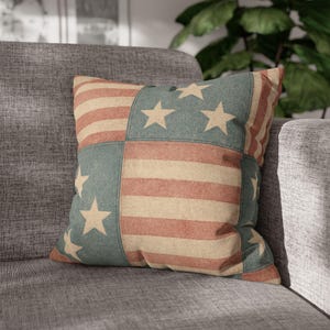 May include: Square throw pillow with a patchwork design featuring red and beige stripes and blue squares with white stars. The pillow is placed on a gray sofa, creating a patriotic theme.