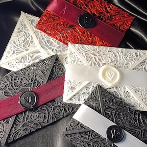 May include: Five rectangular envelopes made of intricate, laser-cut lace in red, white, and black. Each envelope is wrapped with a solid-coloured satin ribbon in red, white, or silver, and secured with a circular wax seal featuring a heart design.