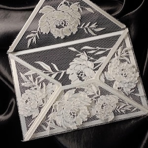 May include: A white lace envelope featuring an intricate openwork mesh pattern and detailed floral embroidery. The design includes several large, textured flower blossoms and delicate leaves integrated into the lace structure, forming a decorative stationery piece.