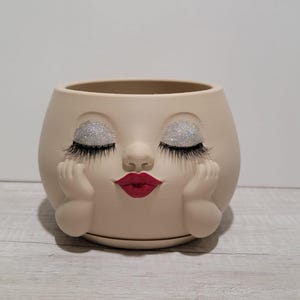 May include: A beige planter shaped like a face with closed eyes, silver glitter eyelids, long black eyelashes, and bright red lips. The planter features two small hands resting against the cheeks on either side of the face, creating a resting pose.