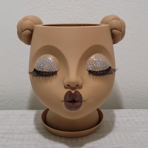May include: A light brown head-shaped planter featuring two braided buns on the sides, closed eyes with silver glitter eyeshadow, long black eyelashes, and dark brown lips. The planter includes a matching circular base tray for drainage.