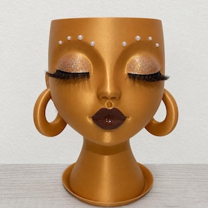 May include: A gold-colored head-shaped planter featuring closed eyes with glittery gold eyeshadow and long black eyelashes. The face is adorned with small white pearl-like accents above the eyes, dark brown glossy lips, and large gold hoop earrings.