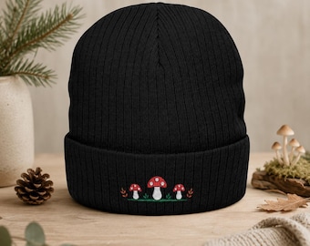 Pode incluir: Um gorro de malha canelada preto com um design bordado de três cogumelos vermelhos com pintas brancas sobre um pequeno pedaço de relva verde, acentuado por pequenas folhas cor de laranja nos lados esquerdo e direito do grupo.