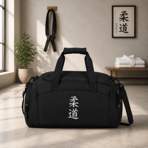 Puede incluir: Una bolsa de deporte negra con dos asas superiores, una correa de hombro extraíble y un compartimento principal con cremallera. La parte frontal de la bolsa muestra los caracteres kanji japoneses blancos para Judo, orientados verticalmente.