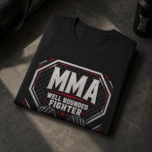 Può includere: Una t-shirt nera con una grafica bianca e rossa di una gabbia ottagonale. Il testo recita MMA WELL ROUNDED FIGHTER, accompagnato da illustrazioni di due guantoni da combattimento neri e tre piccole stelle rosse sotto il testo.