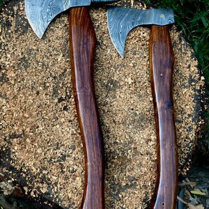 May include: Two axes with patterned steel blades and dark wood handles rest on a wood surface covered in wood shavings. The axes have a curved handle design and a sharp blade edge. The blades have a unique pattern.