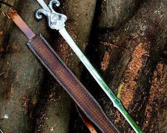 May include: A silver-toned metal sword featuring a fleur-de-lis crossguard and a long, pointed blade. Beside the sword lies a brown, textured leather scabbard with a matching brown leather strap and a metal buckle for carrying or attachment.