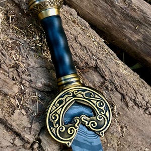 May include: A decorative sword with a gold-coloured hilt and guard, and a black leather-wrapped handle. The blade has a patterned design. The sword is resting on a bed of wood and leaves.