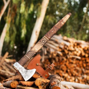 May include: A handcrafted axe with a wooden handle wrapped in leather and decorative carvings. The axe head is silver with intricate designs. A brown leather sheath rests beside the axe. The axe is displayed on a pile of wood.