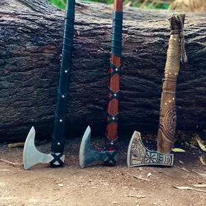 Könnte beinhalten: Drei handgefertigte Äxte mit silbernen Klingen und Holzgriffen. Die Griffe sind mit schwarzem Leder, braunem Holz und Naturfaser umwickelt. Die Axtköpfe weisen aufwendige Designs auf.