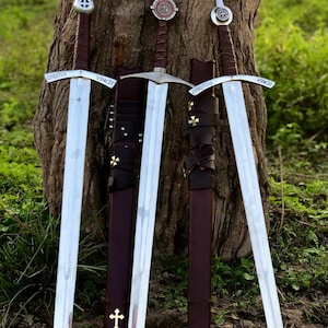 May include: Three medieval swords with silver blades and brown leather-wrapped handles are displayed. Each sword has a decorative cross on the hilt and the words "CHRISTUS VINCIT" engraved on the crossguard. The swords are sheathed in brown leather scabbards.
