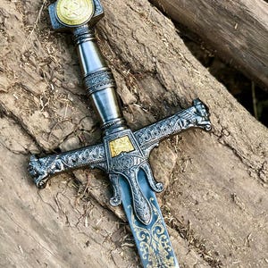 May include: An intricately designed sword with silver and gold detailing. The blade has a blue tint and is adorned with gold filigree. The hilt and crossguard are highly decorated. The pommel features a gold emblem.