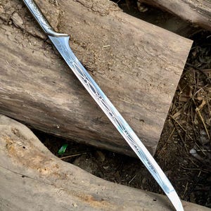 May include: A silver sword with an ornate handle and blade design. The blade has a curved shape with decorative etchings. The sword is resting on a weathered wooden surface, suggesting a fantasy or historical theme.