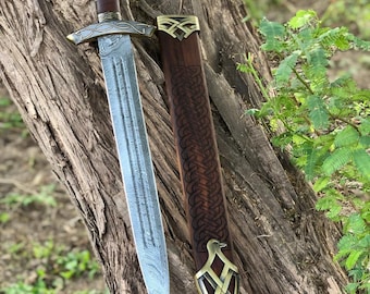 May include: A decorative sword with a silver blade and a brown handle with gold accents. The sword is displayed with its brown leather sheath, which has gold-colored metal detailing. The sword is resting on a tree trunk.