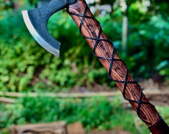 May include: A handcrafted axe with a dark, forged steel blade and a wooden handle. The handle is wrapped with black leather in a crisscross pattern. The axe features etched runes and a leather grip at the base. The background is blurred green foliage.