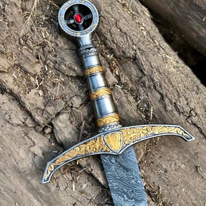 May include: A detailed sword with a silver and gold handle, a patterned blade, and a red gem at the pommel. The crossguard features ornate gold detailing. The sword rests on a wooden surface.