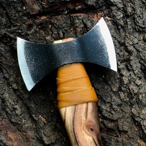 May include: A double-bit axe with a dark, textured blade and a wooden handle. The handle is wrapped with tan leather near the axe head. The axe is set against a dark, textured wood background.