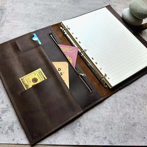 May include: Open brown leather binder with a lined notepad, pockets, and a zippered compartment. A hundred-dollar bill and a credit card are visible in the pockets. A passport is also visible. The binder is open on a gray surface.