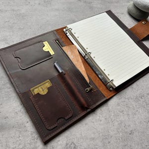 May include: Open brown leather binder with a three-ring mechanism, lined paper, and multiple pockets. Includes a pen, zipper pocket, and card slots. The binder is open, revealing its interior.